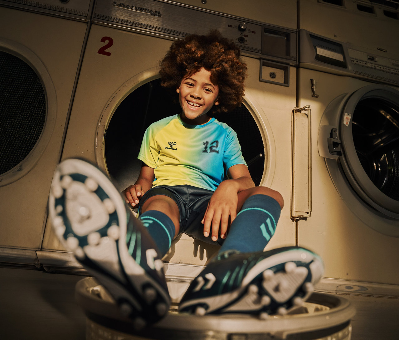 Lächelnder Junge im hummel® Kinder-Trikot-Set HMLSHIMMER, limette, sitzt vor Waschmaschinen.
