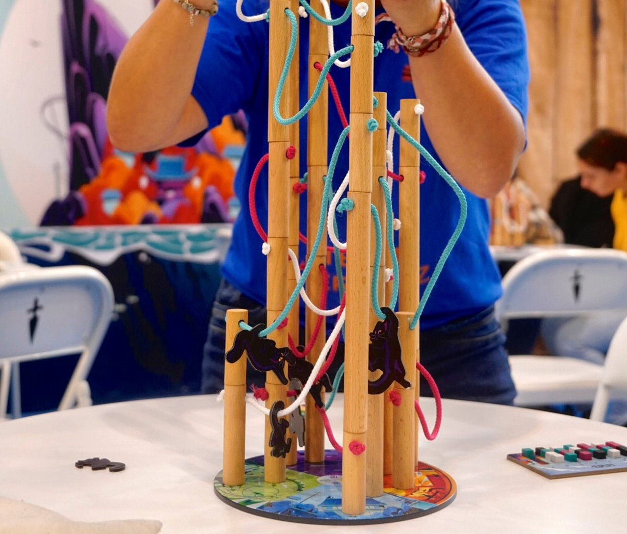 Eine Person spielt mit dem »Nekojima« Spiel, das aus Holzstäben, Schnüren und Katzenfiguren besteht.