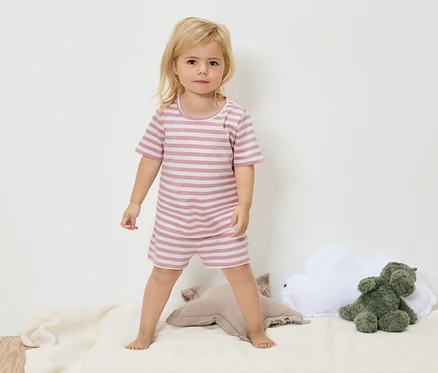 Ein kleines Mädchen mit blonden Haaren steht auf einer cremefarbenen Decke und trägt einen gestreiften rosa-weißen Pyjama.