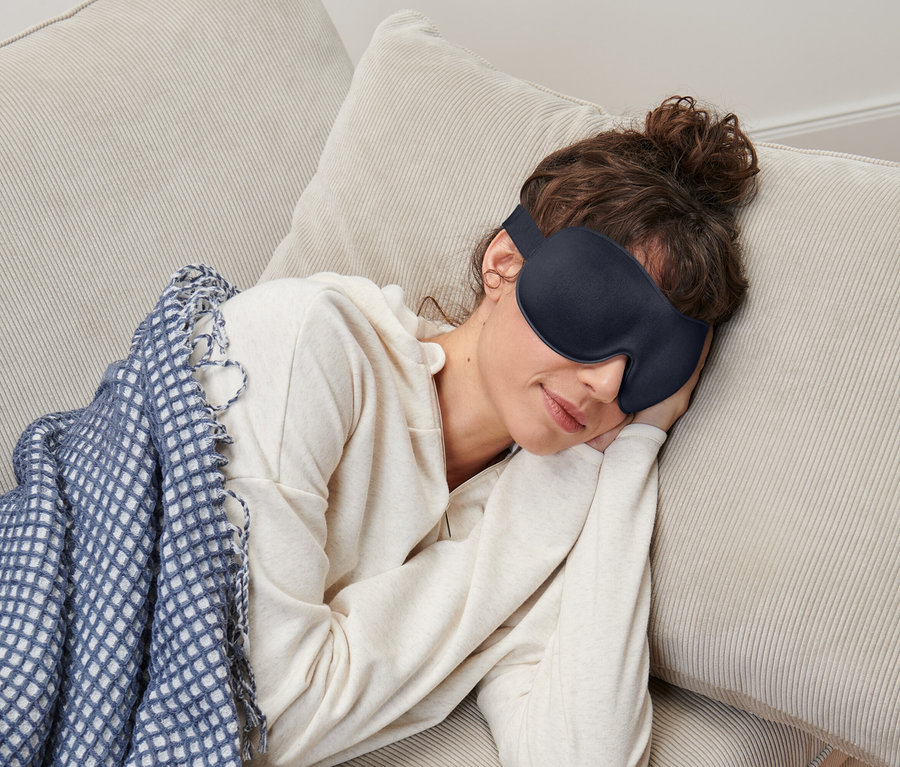 Eine Frau mit hochgesteckten braunen Haaren, die eine Schlafmaske trägt und auf einer beigen Cordcouch unter einer blau-weißen Decke liegt.