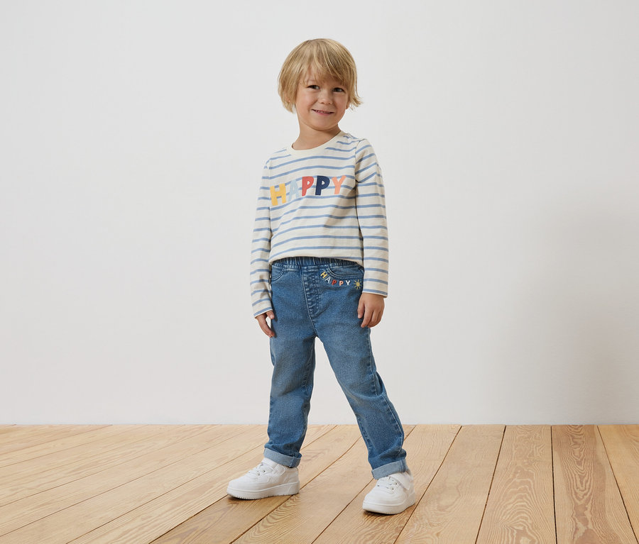 Ein kleiner Junge steht vor einer weißen Wand und trägt ein gestreiftes Langarmshirt mit dem Wort "Happy" und blaue Jeans mit weißen Turnschuhen.