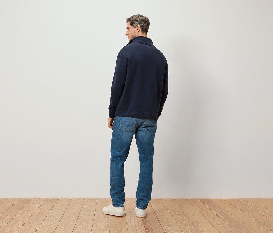 Ein Mann steht mit dem Rücken zur Kamera, er trägt einen marineblauen Pullover mit Reißverschluss, Jeans und weiße Turnschuhe.