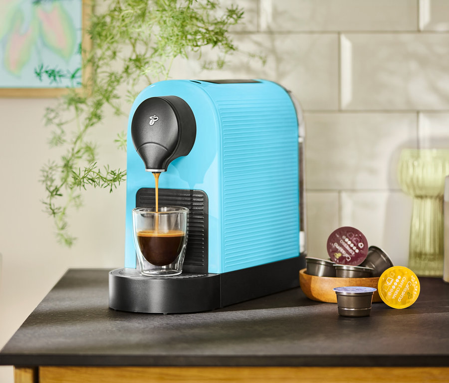 Eine türkisfarbene Kaffeemaschine füllt eine Tasse mit Kaffee. Kaffeekapseln liegen in einer Holzschale daneben.