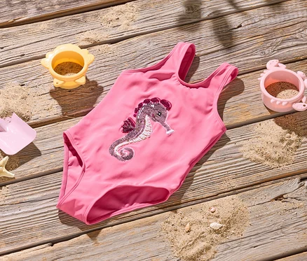 Ein pinkfarbener Badeanzug mit einem Seepferdchen-Design liegt auf einem Holzboden, umgeben von Sand, einer Schaufel, einem Seestern und zwei Sandförmchen.