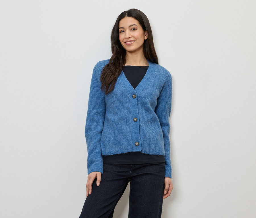 Frau mit dunklen Haaren trägt eine blaue Strickjacke mit drei Knöpfen über einem schwarzen Oberteil und einer dunklen Hose.