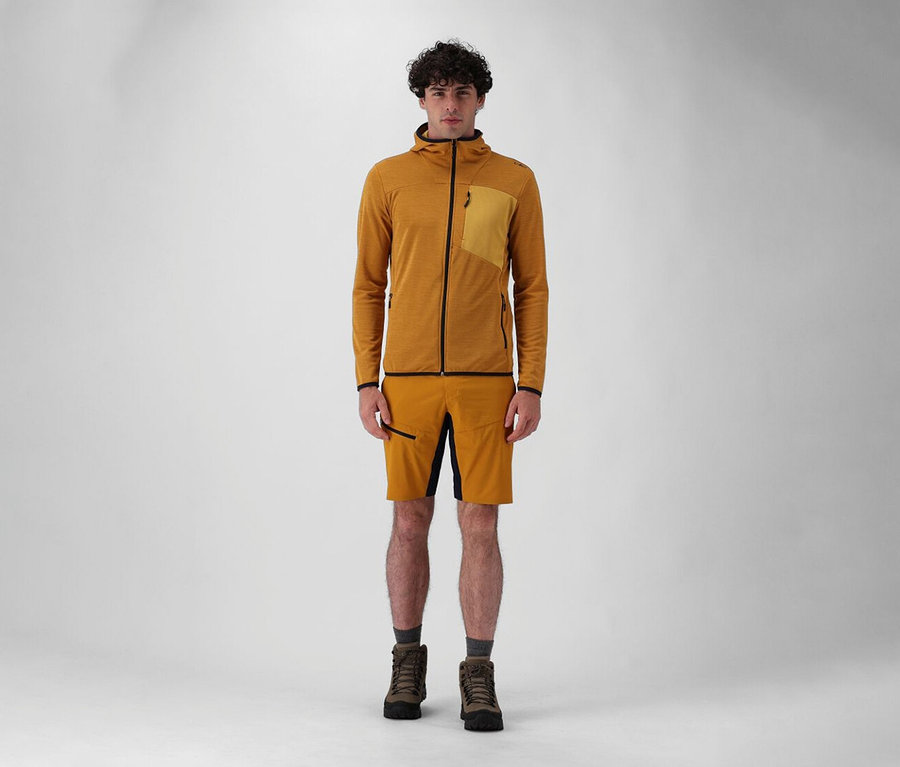 Mann steht in Shorts und Stiefeln mit CMP Herren-Kapuzen-Jacke in Senfgelb vor einem weißen Hintergrund.