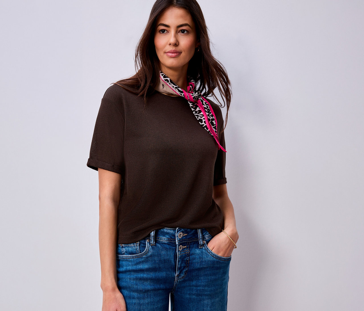 Eine Frau trägt ein dunkelbraunes Street One T-Shirt und blaue Jeans, mit einem pinken Schal mit Animal-Print um den Hals, und hat eine Hand in der Tasche.
