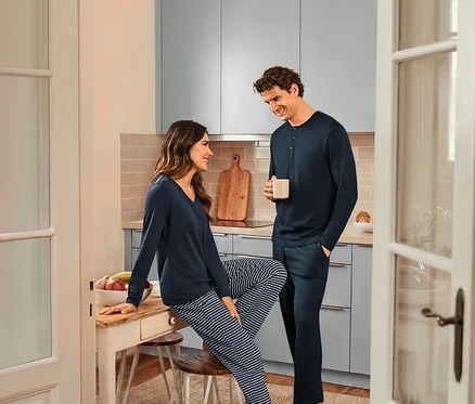 In einer Küche stehen eine Frau, die auf einem Tisch sitzt und einen dunkelblau gestreiften Pyjama und ein dunkelblaues Oberteil trägt, und ein Mann, der eine Tasse hält und einen dunkelblauen Pyjama trägt.