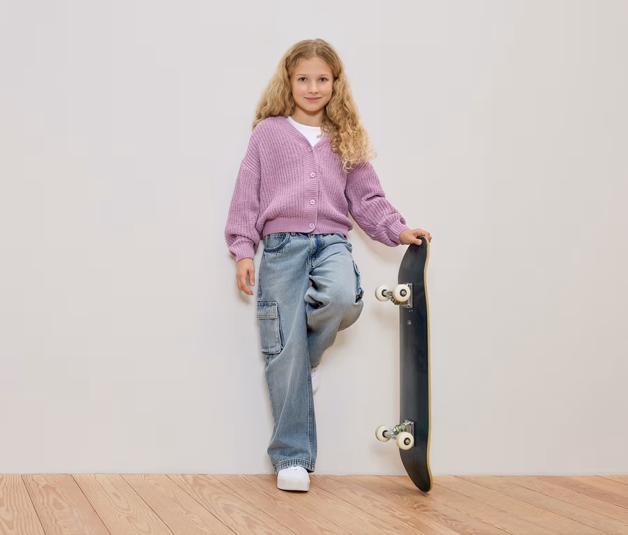 Mädchen mit blonden Locken, trägt einen lila Strickjacke und Jeans, lehnt an einem Skateboard vor einer weißen Wand.