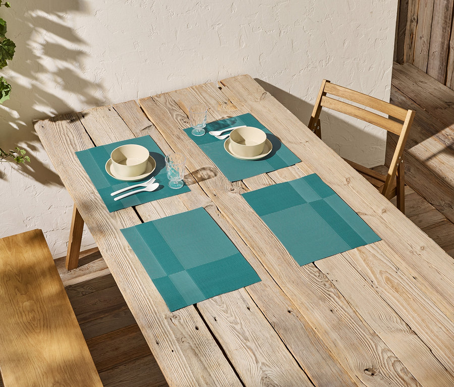 Holzesstisch mit vier türkisfarbenen Tischsets, jeweils mit Schüssel, Teller, Besteck und Glas.