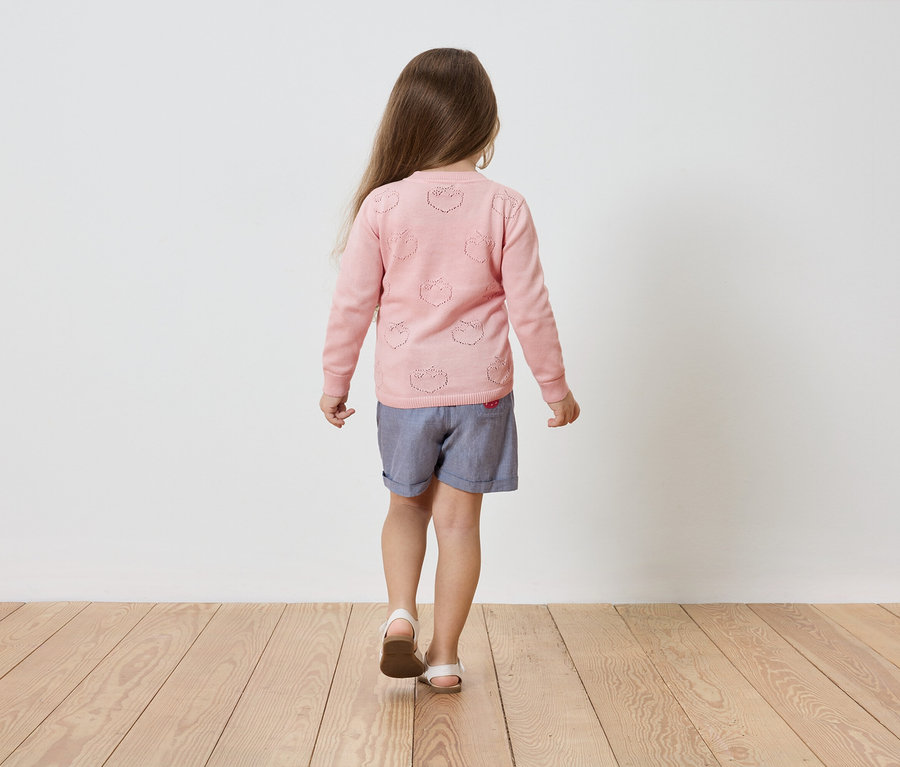 Mädchen von hinten in einem rosa Cardigan mit Erdbeer-Lochmuster, blauen Shorts und Sandalen auf einem Holzboden.
