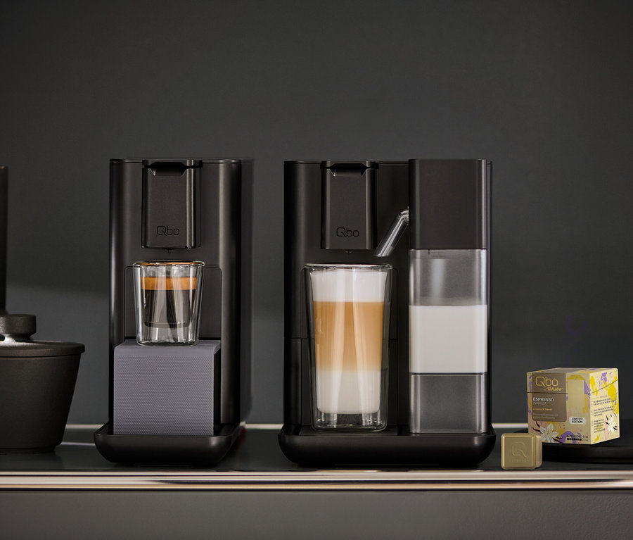 Zwei schwarze Qbo Kaffeemaschinen auf einem dunklen Regal. Die linke Maschine hat ein Glas mit Espresso. Die rechte Qbo MILK MASTER Velvet Black hat ein Glas mit geschichtetem Latte Macchiato und einen Milchbehälter. Rechts daneben stehen eine gelbe Packung Qbo Espresso-Kapseln und eine goldene Kapsel.