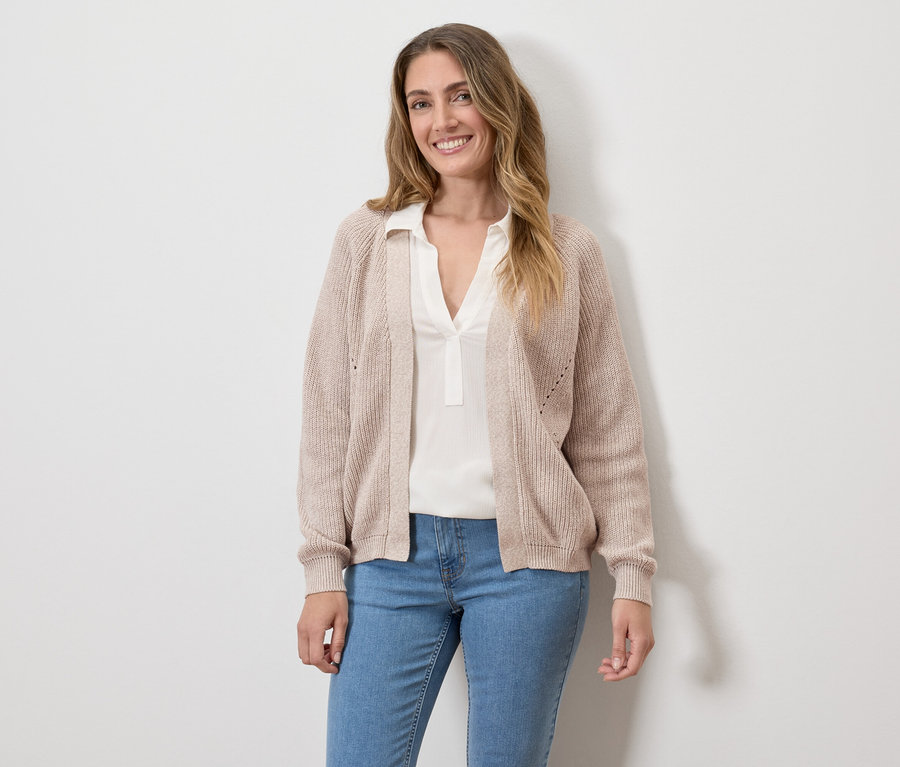 Frau mit beigem Cardigan, weißer Bluse und Jeans vor einer weißen Wand.
