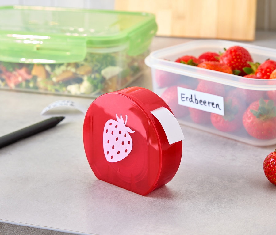 Ein roter Etikettenspender mit Erdbeermotiv steht neben einer beschrifteten Erdbeerbox und einer Salatbox auf einer grauen Oberfläche.