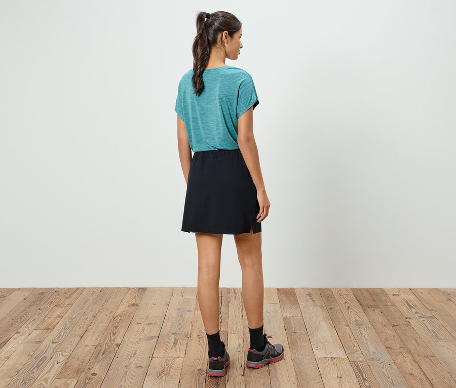 Eine Frau steht mit dem Rücken zur Kamera, trägt ein blaugrünes T-Shirt, einen schwarzen Rock und Sportschuhe.