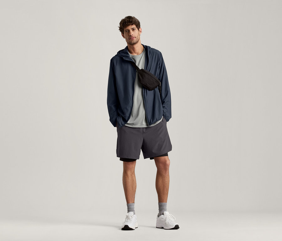 Mann steht und trägt ein hellgrau meliertes Sportshirt, eine marineblaue Jacke und graue Shorts.