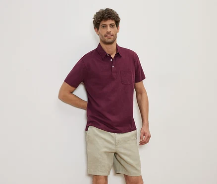 Ein Mann lehnt an einer Wand und trägt ein weinrotes Poloshirt und beige Shorts.