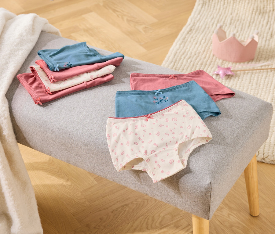 Auf einer grauen Bank liegen gefaltete Mädchenslips in Rosa, Weiß und Blau, daneben eine Spielzeugkrone und ein Zauberstab.