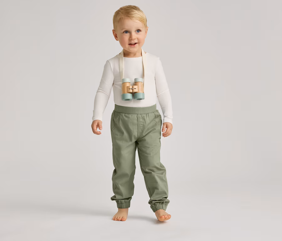 Ein barfüßiger Junge mit blauen Augen und blonden Haaren steht mit einer hellen Hose und einem hellen Oberteil vor einem grauen Hintergrund.