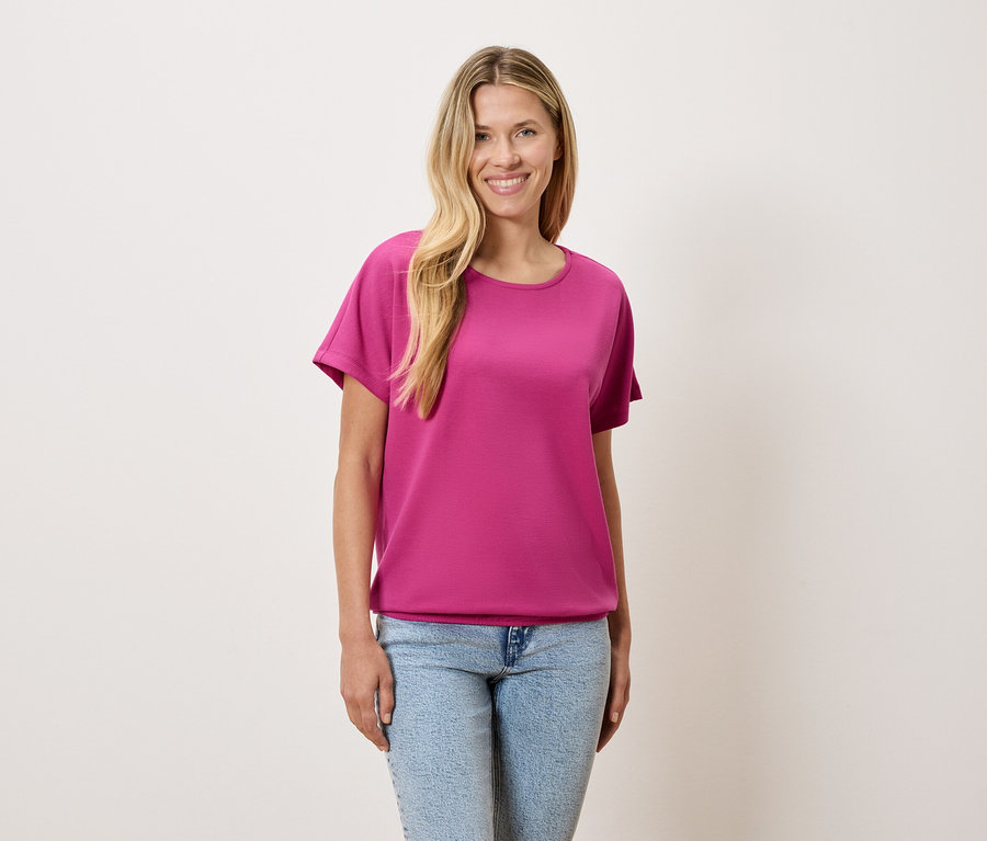 Eine Frau mit blonden Haaren trägt ein pinkes T-Shirt und blaue Jeans.