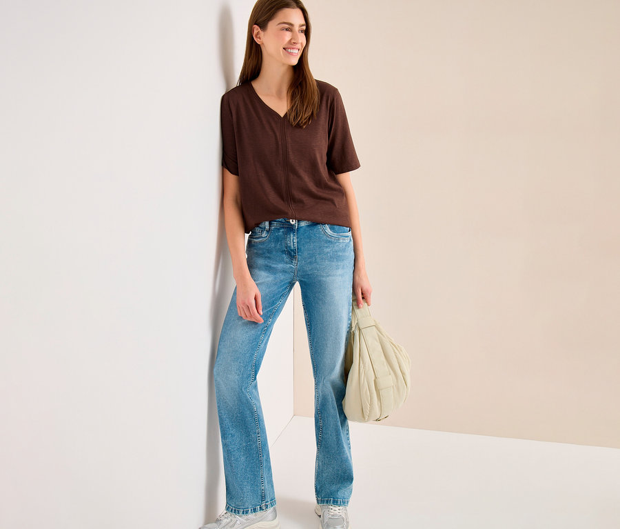 Eine lächelnde Frau trägt ein braunes CECIL Tunika-Shirt, hellblaue Schlagjeans und silberne Sneaker, lehnt an einer weißen Wand und hält eine hellbeige gesteppte Handtasche.
