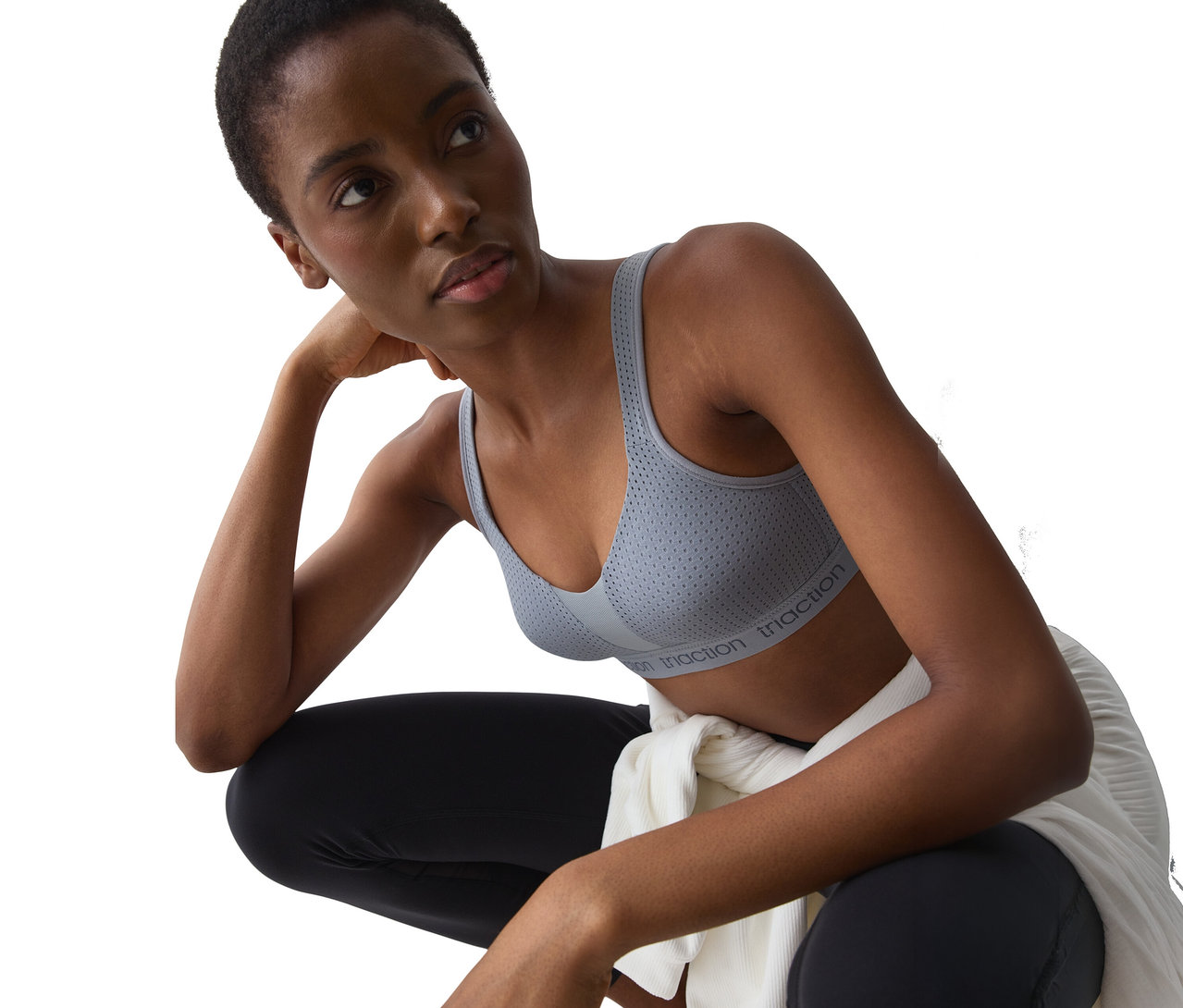 Eine Frau mit kurzen dunklen Haaren, die einen grauen, perforierten Sport-BH mit "triaction"-Aufschrift und schwarze Leggings trägt, hockt und blickt nach oben. Ein weißes Kleidungsstück ist um ihre Taille gebunden.