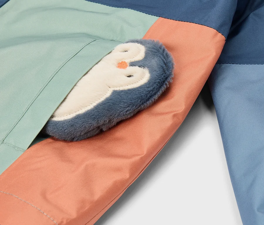 Nahaufnahme eines farblich abgesetzten Kleidungsstücks in Dunkelblau, Hellgrün und Pfirsich, mit einem plüschigen blau-weißen Pinguin mit orangefarbenem Schnabel, der teilweise in einer Naht steckt.