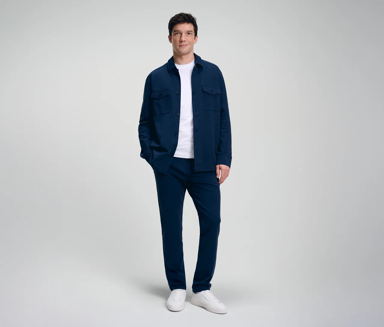 Mann steht, trägt ein dunkelblaues Overshirt über einem weißen T-Shirt, dunkelblaue Hose und weiße Sneaker. Er hat eine Hand in der Tasche und blickt nach vorne.
