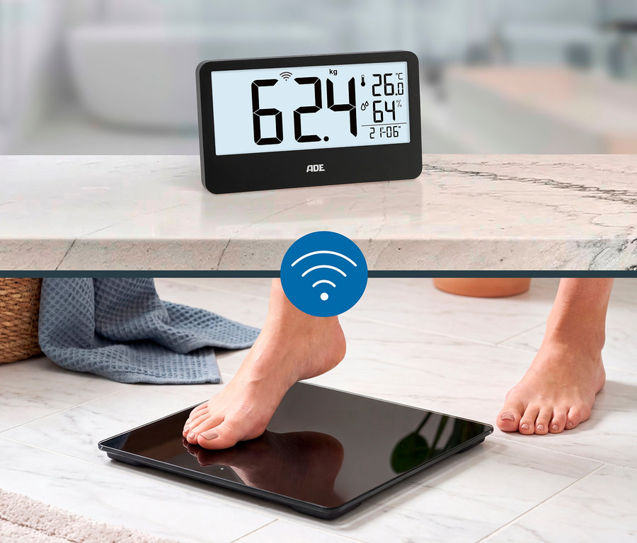 Eine Person tritt mit einem Fuß auf eine schwarze digitale Personenwaage. Ein separates schwarzes Display auf einer Marmorplatte zeigt 62,4 kg, Temperatur, Luftfeuchtigkeit und Uhrzeit an, verbunden durch ein WLAN-Symbol.