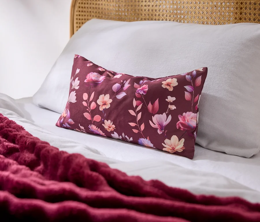 Ein Bett mit weißen Laken, einem weißen Kissen und einem rechteckigen burgunderroten Kissen mit Blumenmuster. Eine strukturierte burgunderrote Decke liegt auf dem Bett, ein Rattan-Kopfteil ist sichtbar.