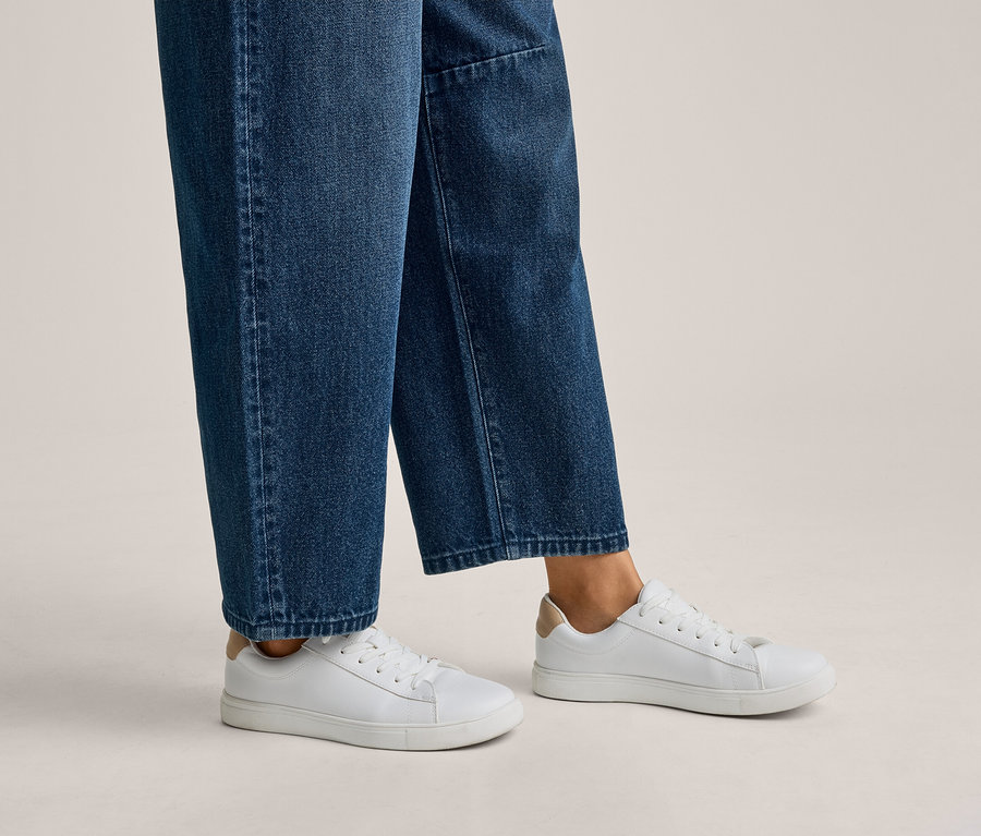 Person trägt blaue Jeans und weiße Sneaker mit beigen Fersenakzenten.