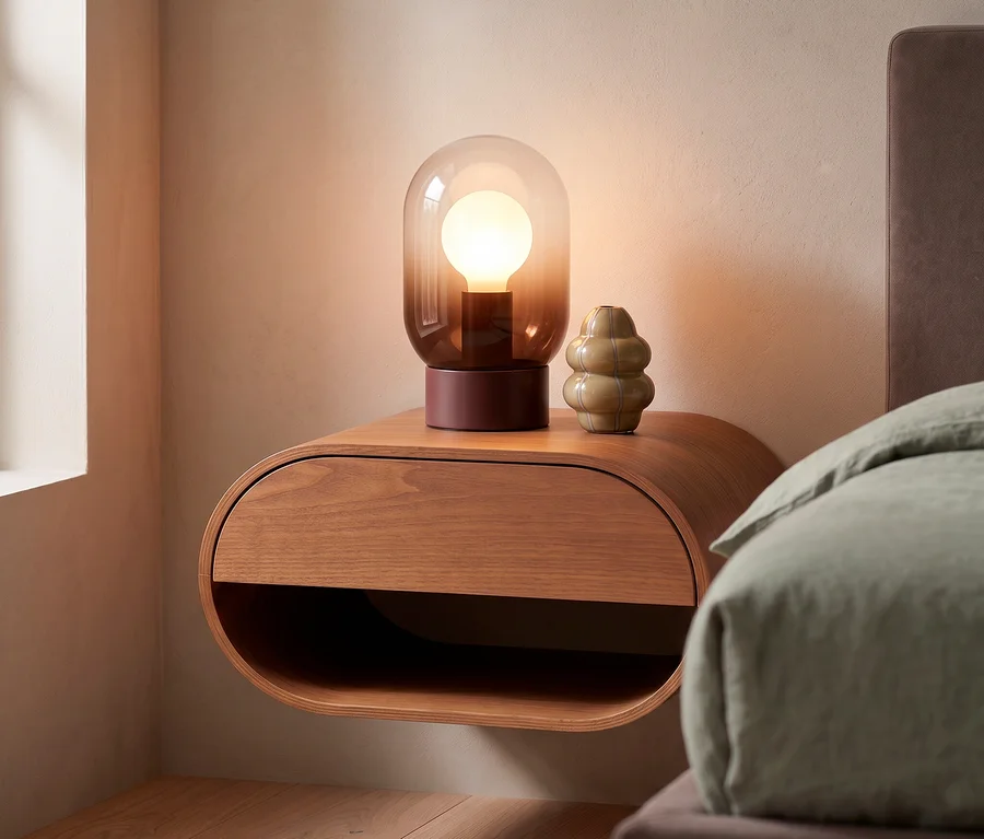 Ein brauner, hängender WOOOD Holz-Nachttisch »Momo« mit einer Lampe und einer grün-grauen WOOOD Glasvase »Kency«. Rechts ist ein Bett mit grünem Bettzeug zu sehen.