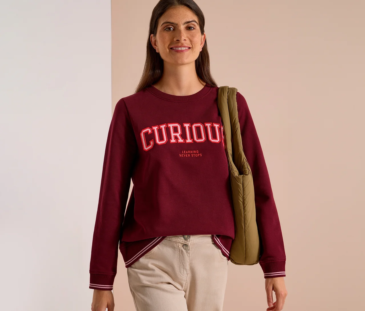 Lächelnde Frau in einem bordeauxroten CECIL Sweatshirt mit der Aufschrift "CURIOUS LEARNING NEVER STOPS" und beigefarbener Hose, eine grüne Tasche über der Schulter tragend.