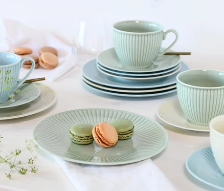 Ein Tisch gedeckt mit pastellgrünem und -blauem creaTable Kombiservice »Valencia Pastell«, bestehend aus geriffelten Tassen, Tellern und Untertassen. Zwei grüne und ein orangefarbener Macaron liegen auf einem pastellgrünen Teller im Vordergrund.