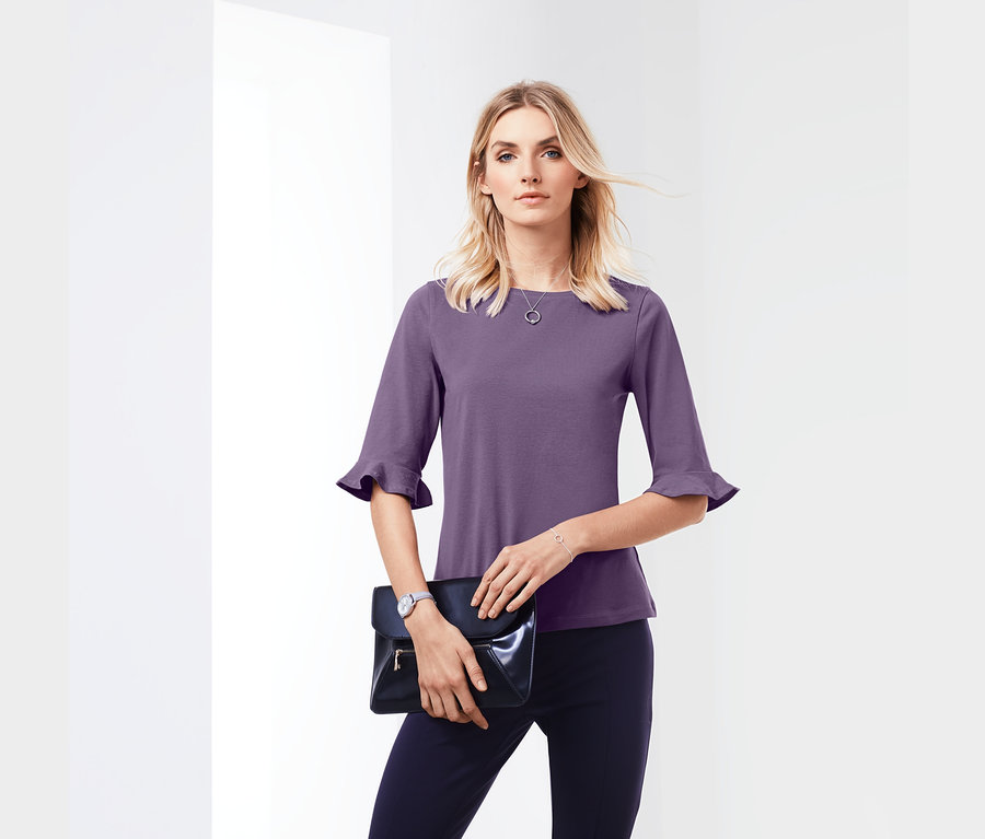 Blonde Frau trägt ein Shirt mit Volants und hält eine schwarze Clutch.