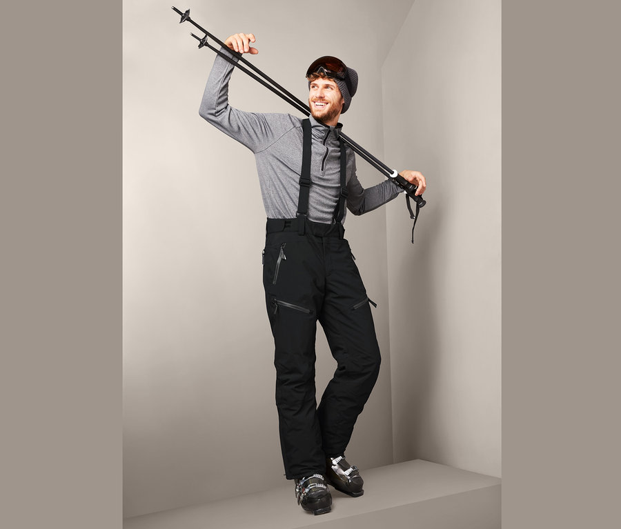 Mann posiert mit Skistöcken über der Schulter, trägt eine schwarze High-End-Skihose und Skischuhe.