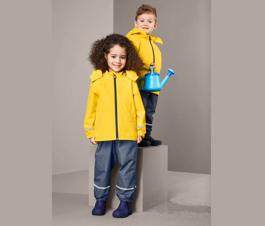 Zwei Kinder posieren: Mädchen in Thermo-Regenjacke aus recyceltem Material und Junge, der eine blaue Gießkanne hält.