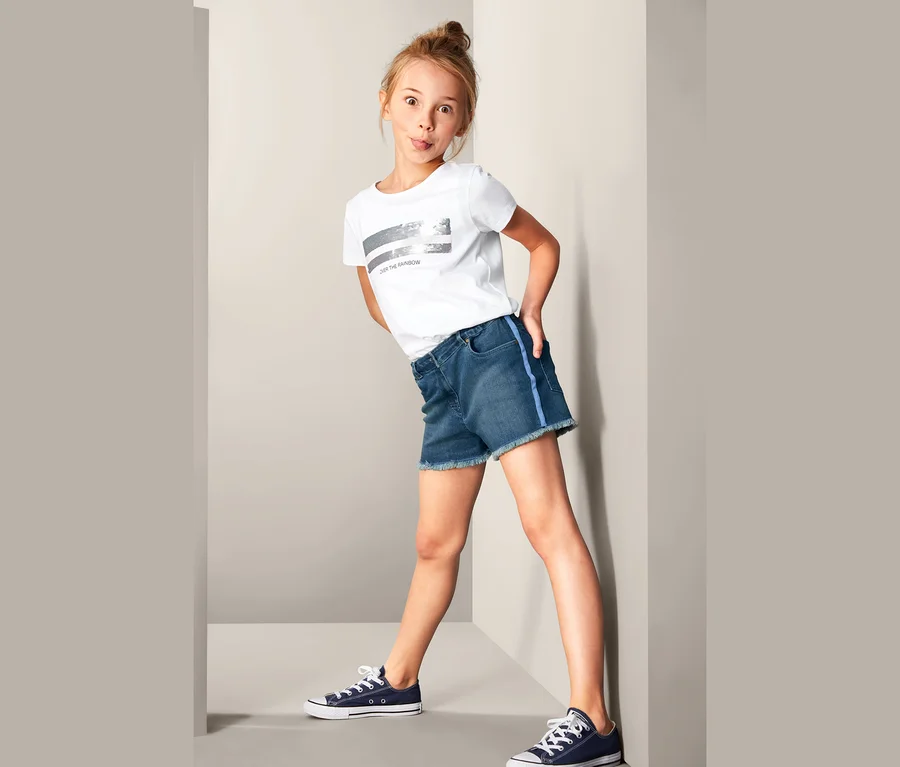 Mädchen posiert in Denim-Shorts, T-Shirt und Turnschuhen.