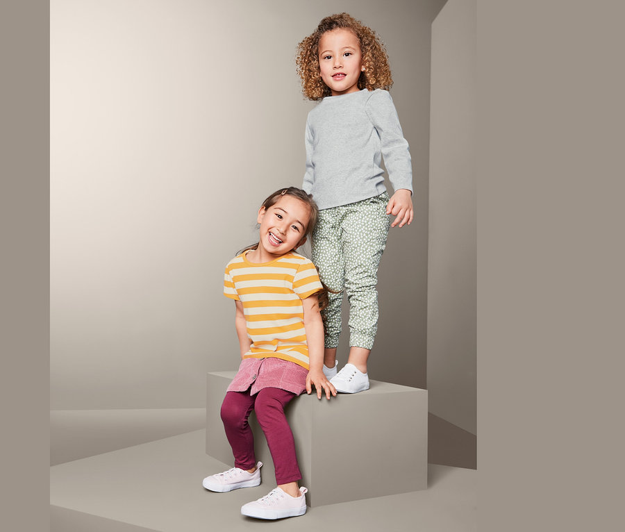 Zwei Mädchen posieren vor einem grauen Hintergrund. Ein Mädchen steht und trägt ein graues 2 Rippenshirts und eine grüne Hose mit Aufdruck. Das andere Mädchen sitzt und trägt ein gelb gestreiftes Shirt und einen rosa Rock.