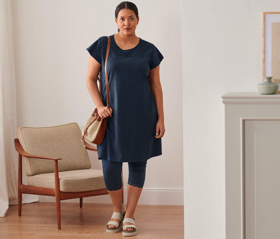Frau steht und trägt ein marineblaues Jerseykleid mit Lochstickerei, 3/4-Leggings mit Spitzenborte, Plateau-Sandale aus Leder und eine Beuteltasche.