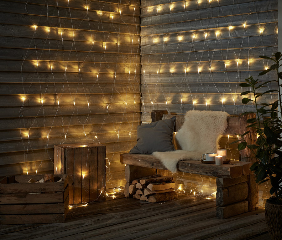 Eine Holzbank mit Kissen und Fell steht vor einer Wand, die mit einem LED-Outdoor-Lichtnetz geschmückt ist.