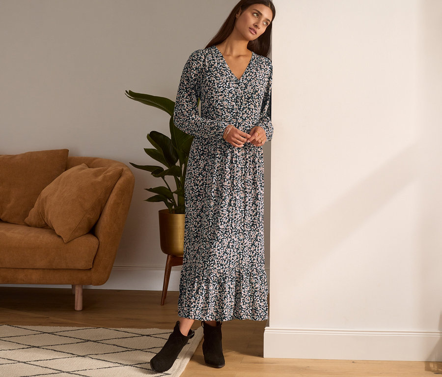 Eine Frau lehnt an einer Wand und trägt ein Maxi-Kleid mit Blumenmuster und schwarze Stiefeletten.