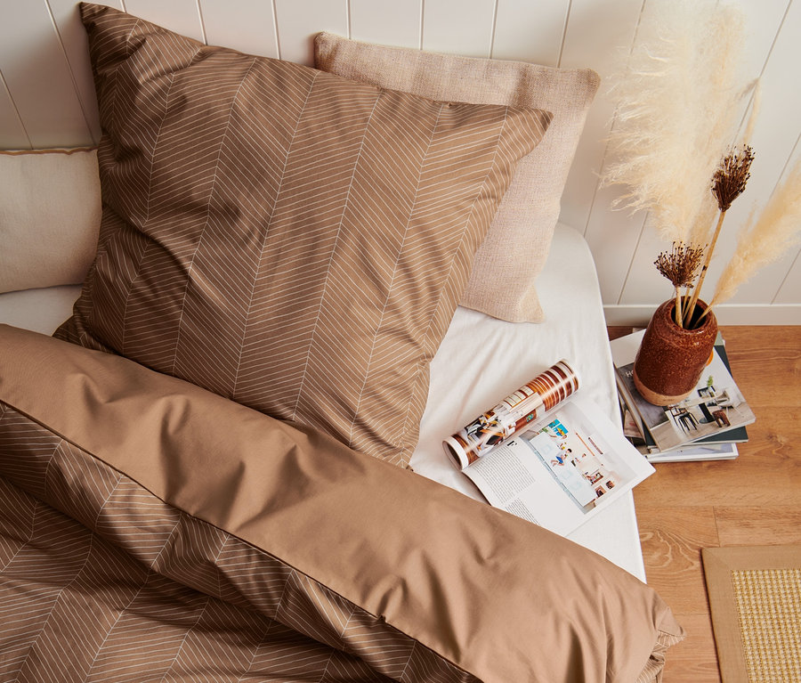 Draufsicht auf ein Bett mit beiger Premium-Baumwoll-Bettwäsche mit geometrischem Muster, daneben eine Zeitschrift und eine Vase mit Pampasgras.