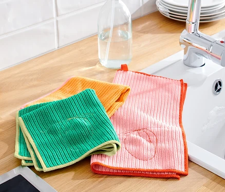 Auf einer Küchentheke liegen grüne, orangefarbene und rosa 3 Universal-Reinigungstücher neben einer Spüle und Ölflaschen.