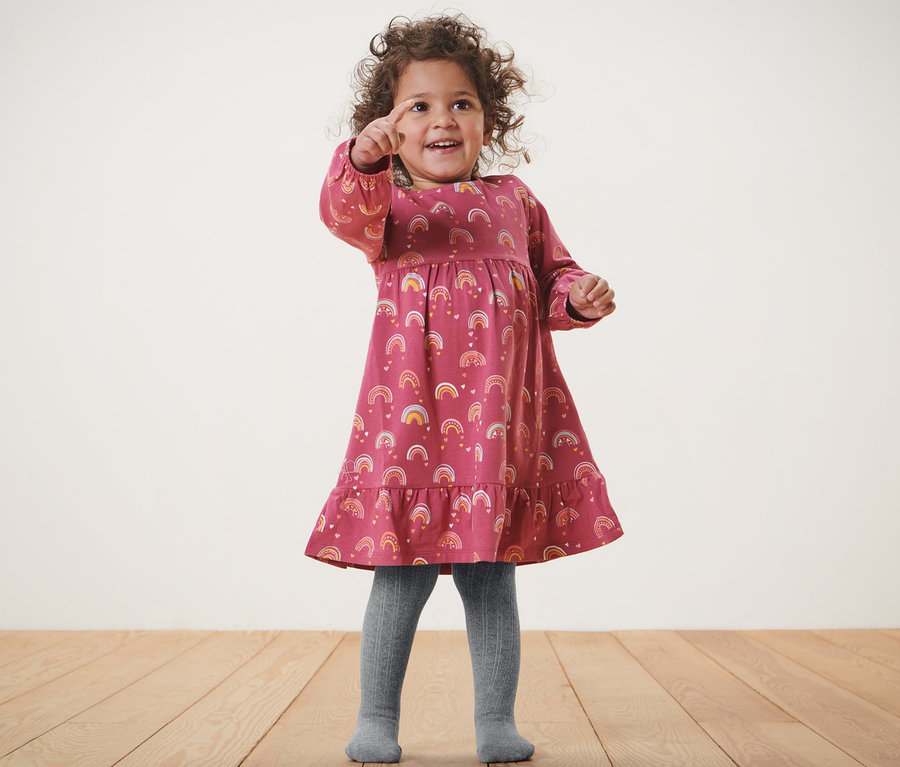 Mädchen im Kleid mit Regenbogenmuster und grauer Baby-Strumpfhose, 2 Paar, zeigt mit dem Finger.