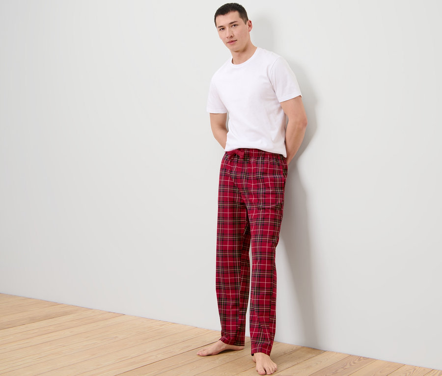 Mann im weißen T-Shirt und Relaxhose mit Karomuster, der an einer Wand lehnt.