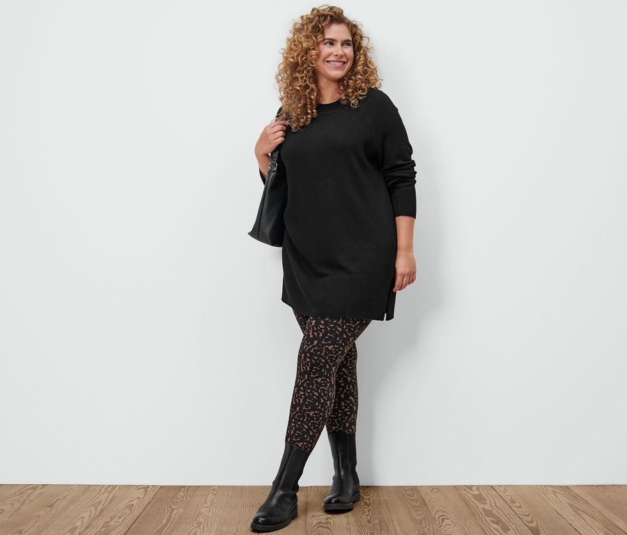 Frau posiert in einem schwarzen, langen Strickpullover, bedruckten Leggings, Lederboots und mit einer Umhängetasche.