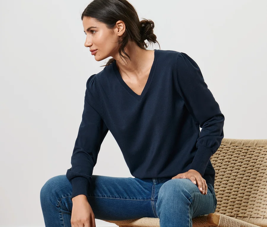 Frau sitzt auf einem Stuhl und trägt einen blauen Feinstrickpullover und Jeans.