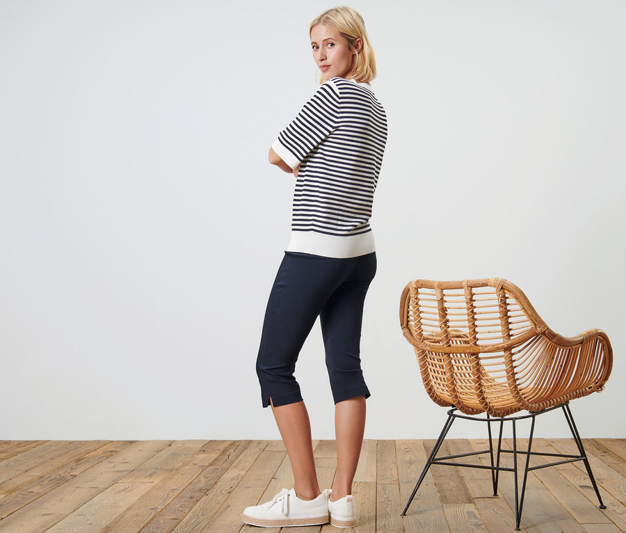 Frau posiert in dunkelblauer 3/4-Stretchhose und gestreiftem Oberteil neben einem Korbstuhl.