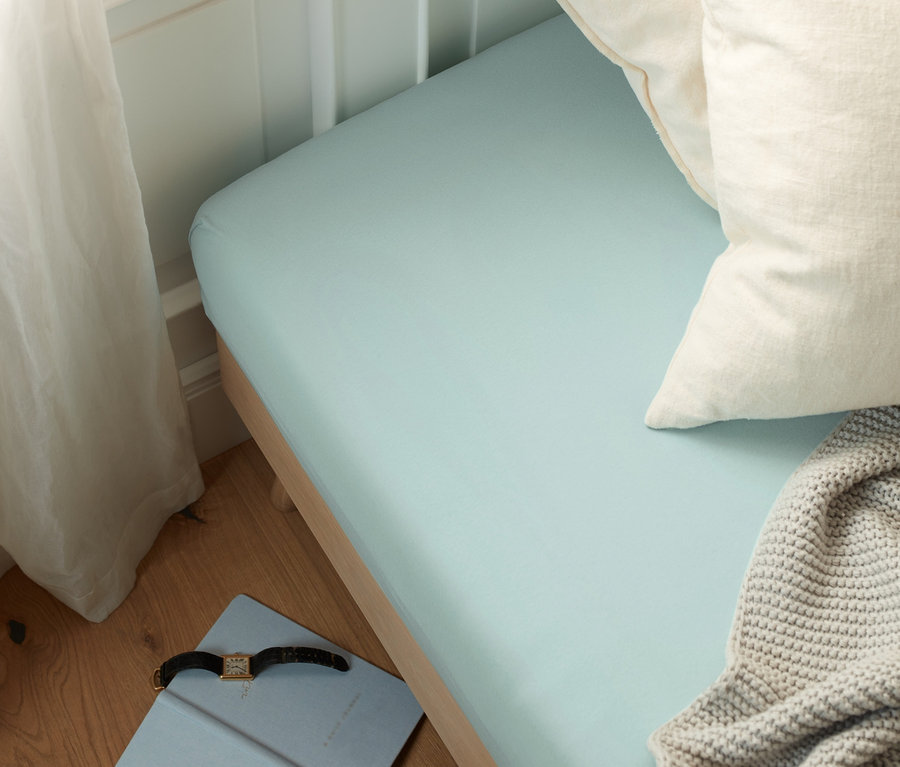 Hellblaues Jersey-Spannbettlaken auf einem Bett mit weißen Kissen und einer grauen Decke. Auf dem Boden liegen ein blaues Notizbuch und eine Uhr.